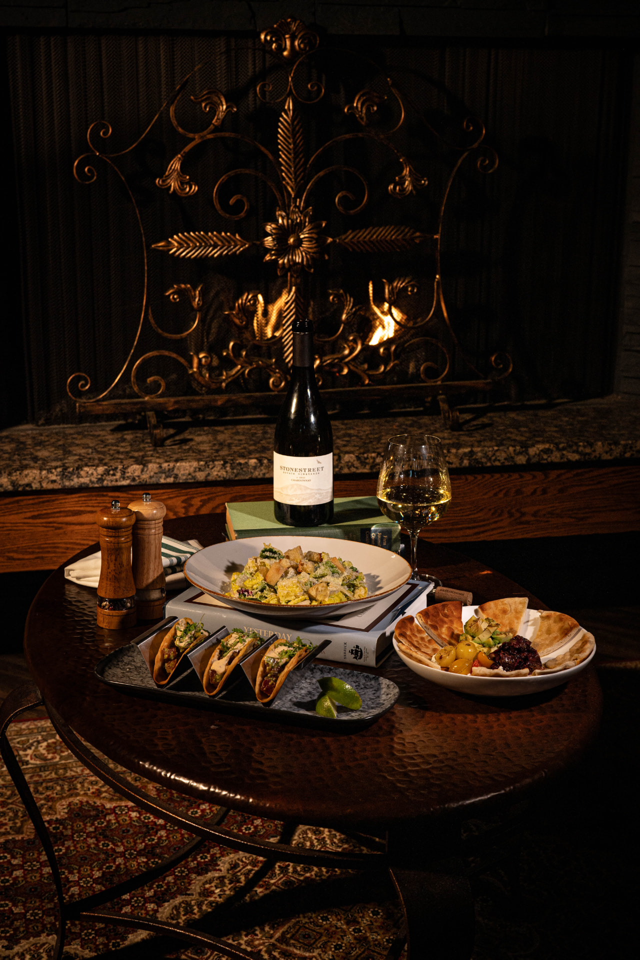 Firelit table set with wine, tacos, salads and dips in front of a fireplace